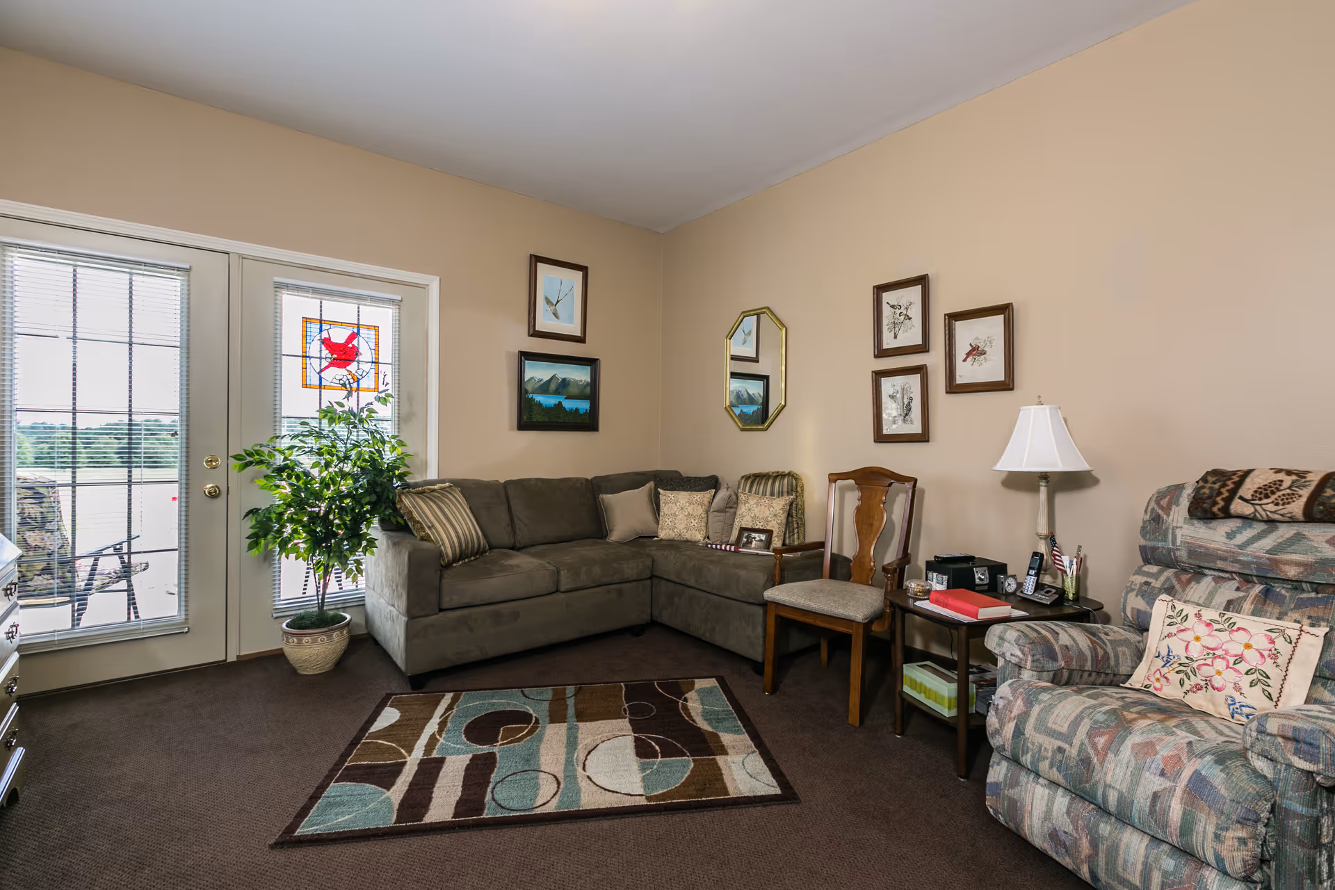 A cozy living room with a gray sectional sofa adorned with pillows, a patterned recliner chair with a floral pillow, a wooden chair, and a small side table with a lamp, phone, and books. The room has beige walls decorated with framed artwork and a mirror. There is a colorful area rug on the carpeted floor and a potted plant near glass doors with blinds and a stained glass window panel.