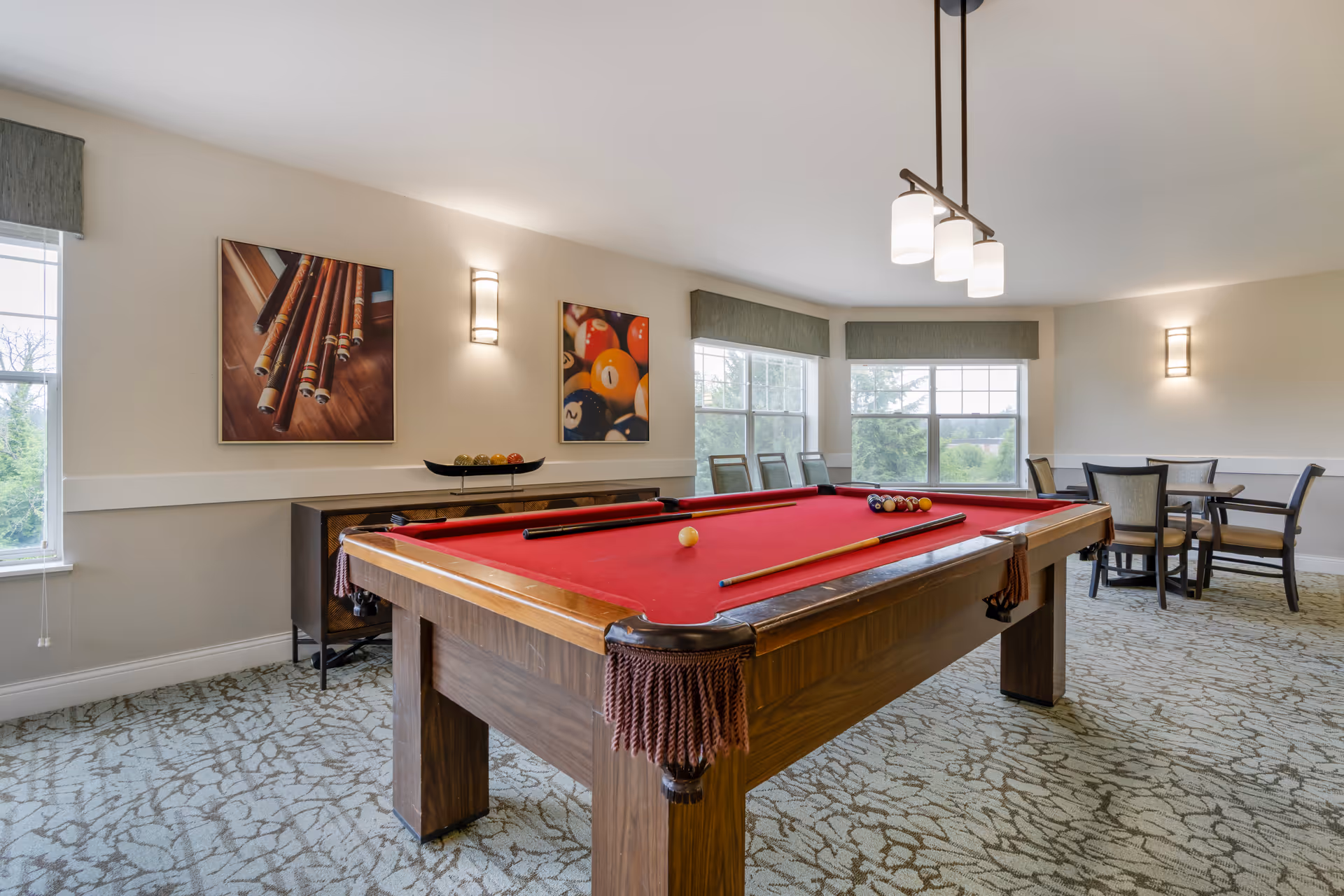 A bright room with a red felt pool table in the center, two pool cues resting on it, and a set of billiard balls arranged for a game. The room has large windows with green valances letting in natural light, patterned carpet flooring, and a small seating area with a table and four chairs in the background. Two framed pictures of billiard balls and pool cues hang on the wall above a wooden sideboard.