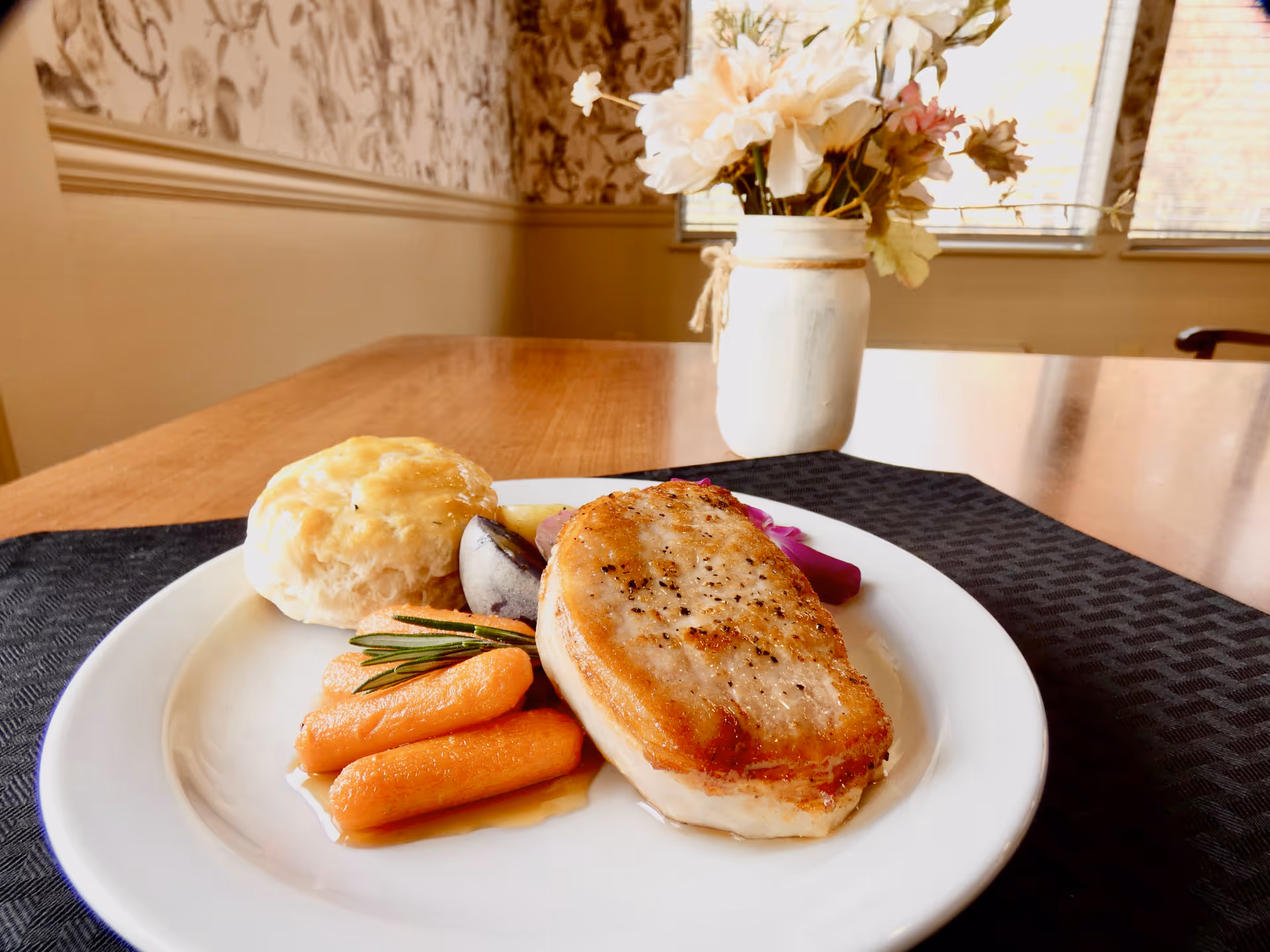 Plate with a seared pork chop, glazed baby carrots and a biscuit on a table with a vase of flowers in the background.