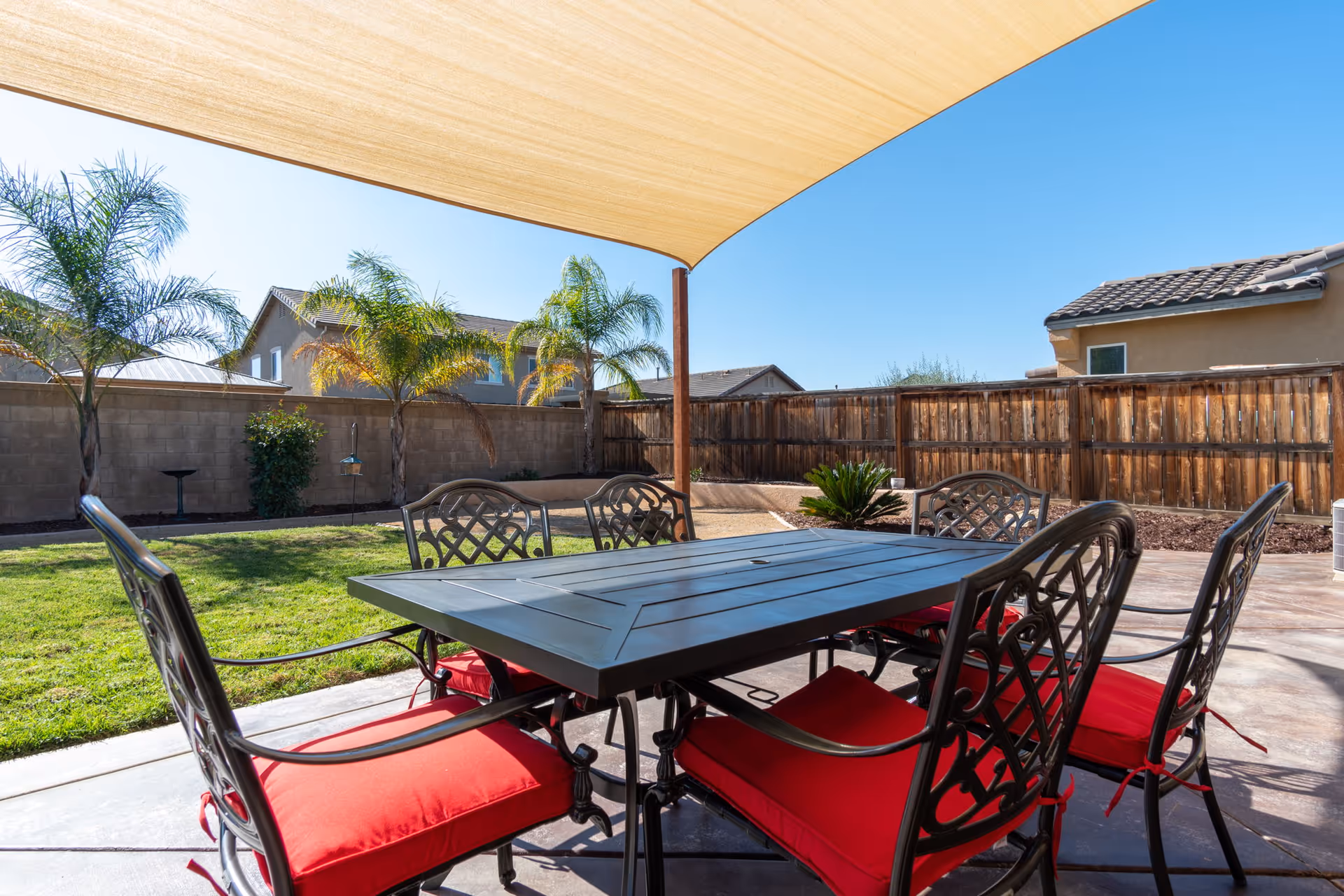 Outdoor patio area with a black metal table and six chairs with red cushions under a beige shade sail. The patio overlooks a grassy yard with palm trees and is enclosed by a wooden fence. Neighboring houses are visible in the background under a clear blue sky.
