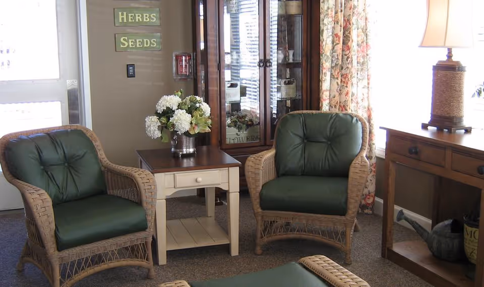 A cozy sitting area with two wicker armchairs featuring dark green cushions, a small wooden side table with a vase of white flowers, a wooden cabinet with glass doors, floral curtains, and a wooden desk with a lamp and a watering can underneath.