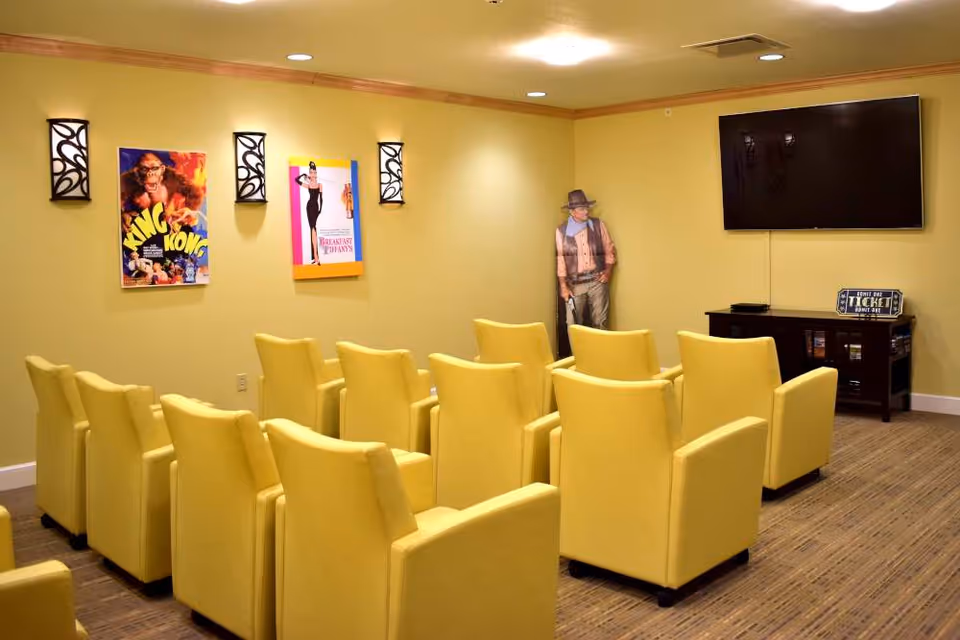 A small movie theater room with two rows of yellow cushioned chairs facing a wall-mounted flat screen TV. The walls are painted light yellow and decorated with movie posters of King Kong and Breakfast at Tiffany's, as well as two decorative wall sconces. A life-sized cardboard cutout of a cowboy stands in the corner next to a wooden media console holding DVDs and a sign that reads 'Admit One Ticket'.