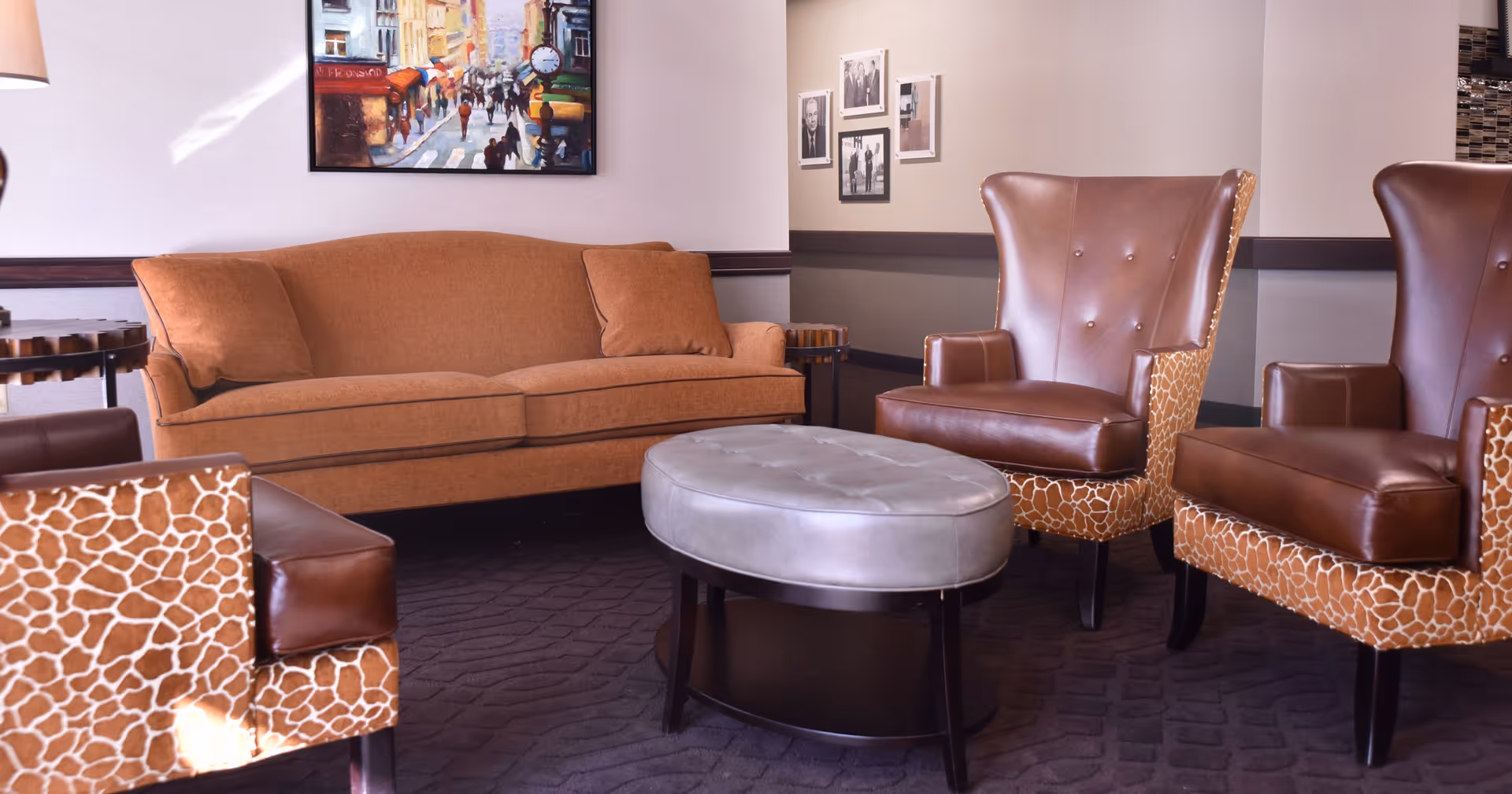 A cozy seating area featuring a brown sofa with two matching cushions, two leather armchairs with giraffe-patterned sides, and an oval gray ottoman in the center. The room has framed artwork on the walls and a dark carpeted floor.