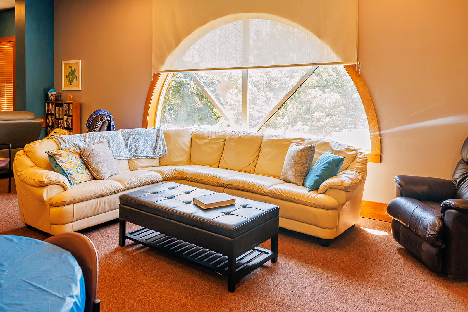 A cozy living room area with a large cream-colored sectional sofa adorned with various pillows and a blue throw blanket. In front of the sofa is a black tufted ottoman with a book on top. To the right is a dark leather recliner chair. The room features a large, arched window with a white roller shade partially drawn, allowing natural light to brighten the space. The walls are painted in warm tones, and there is a small bookshelf and framed artwork visible in the background.