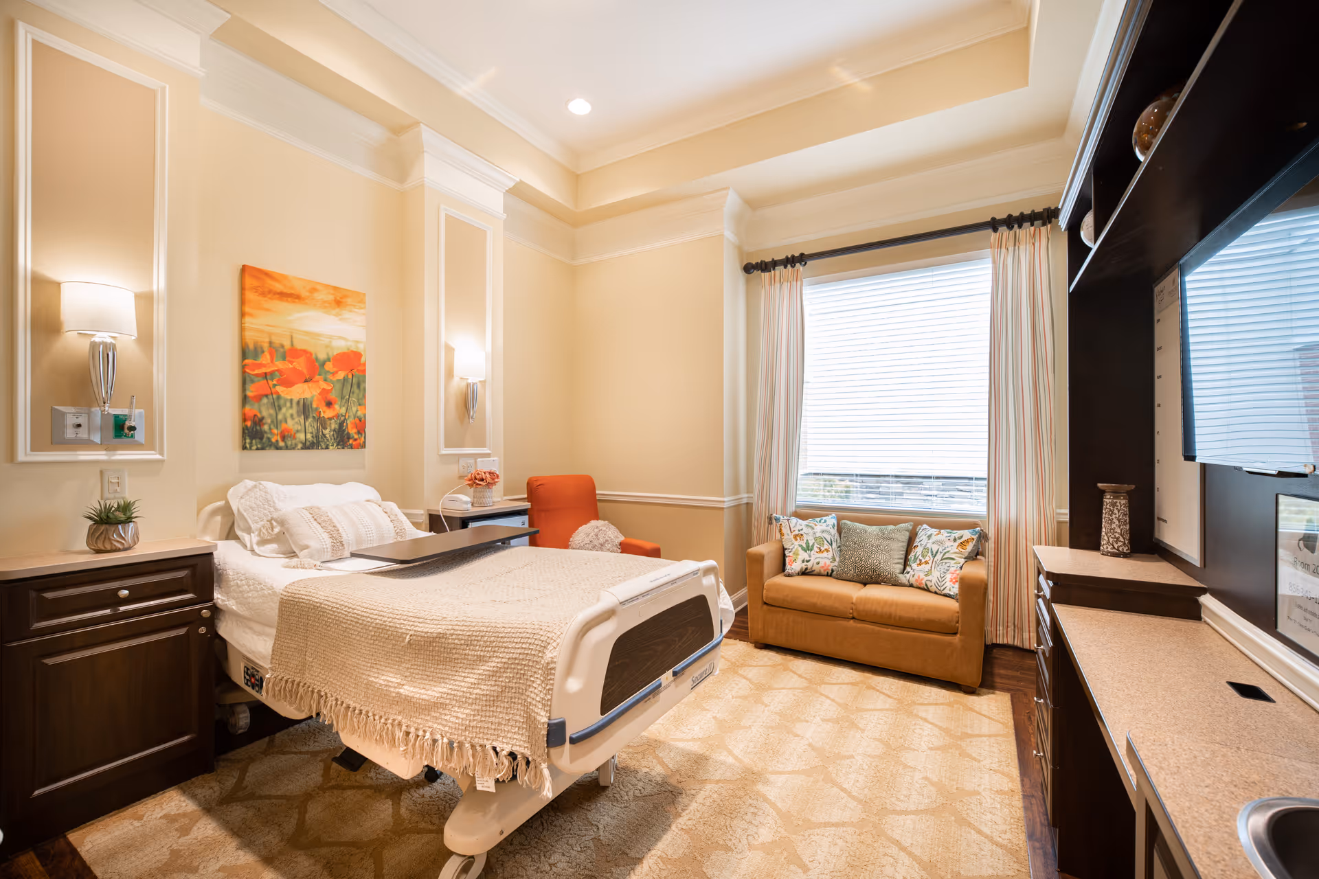 A bright senior-care bedroom with a hospital-style bed, nightstands, built-in cabinetry, a small sofa by a large window, and decorative artwork.