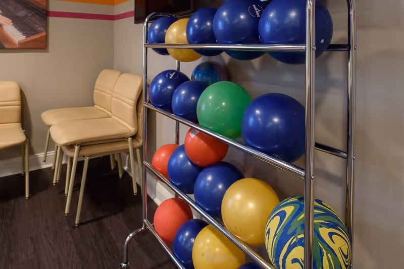 A metal rack holding colorful exercise and medicine balls next to stacked beige chairs in an indoor room.