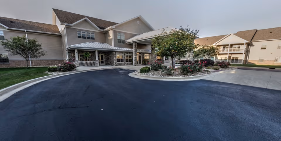 Exterior view of Lincolnshire Place of Decatur showing a large building with beige siding and stone accents. The entrance has a covered drop-off area supported by stone columns. There are landscaped areas with trees and flowering bushes around the driveway and parking area.
