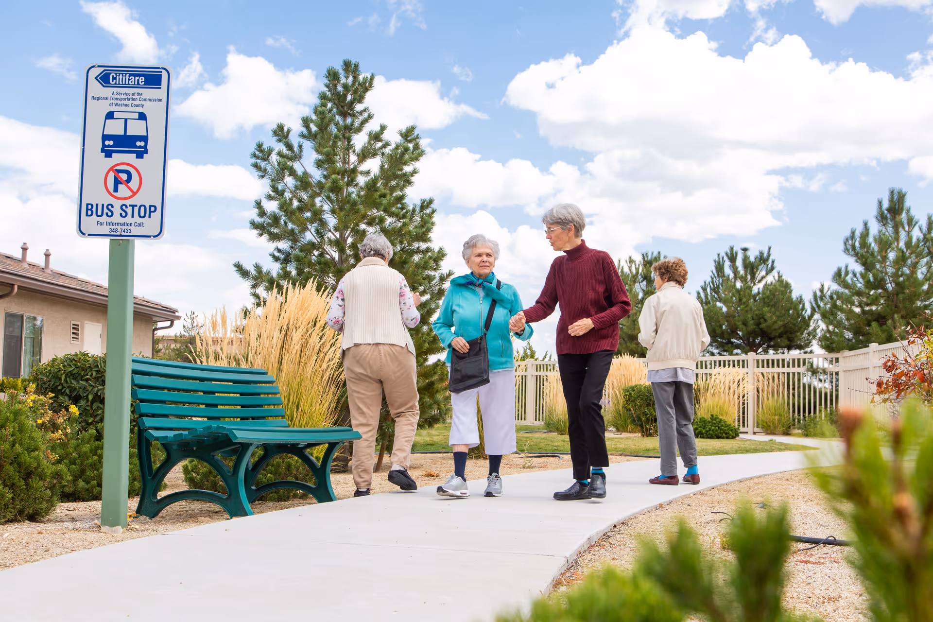 Four elderly women walking and standing on a paved pathway near a green bench and a bus stop sign in an outdoor garden area with trees, bushes, and ornamental grasses under a partly cloudy sky.