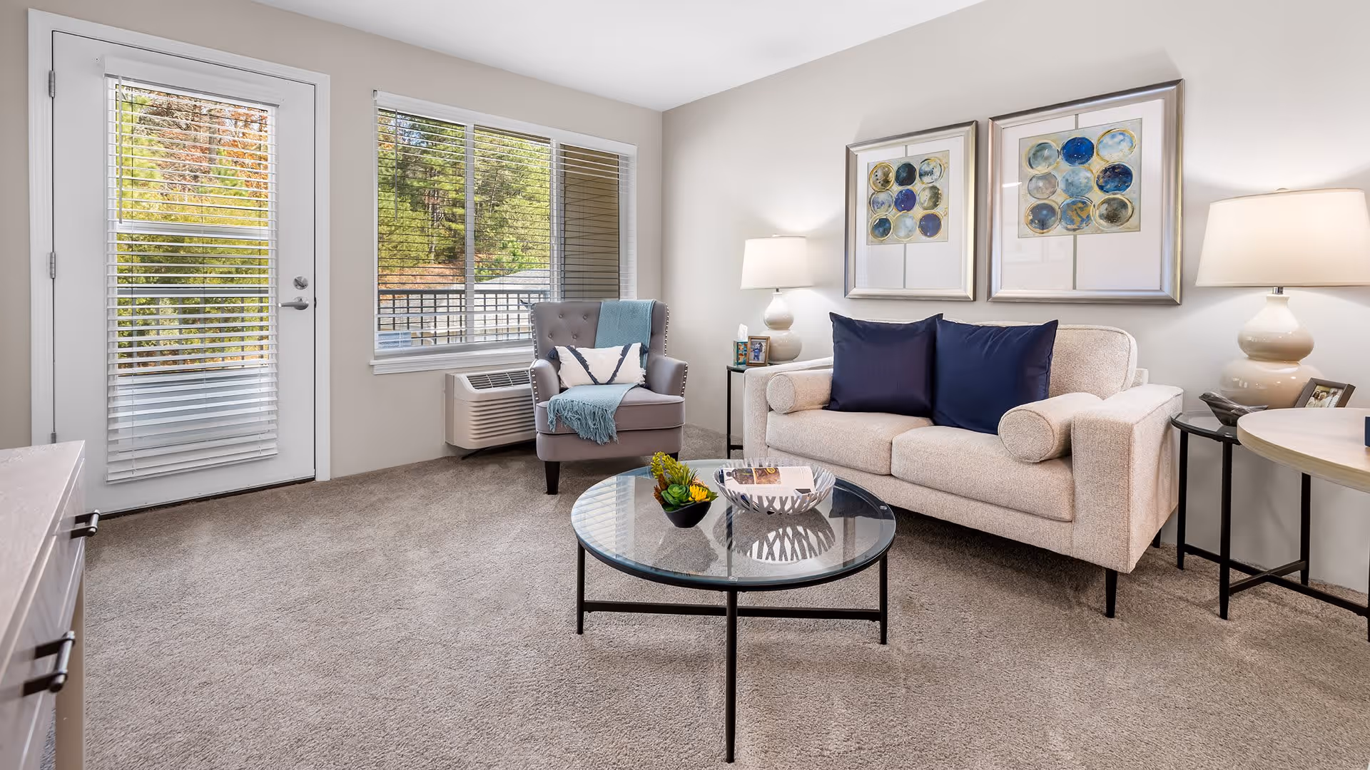 A bright and cozy living room with a beige sofa adorned with two dark blue pillows, a gray armchair with a white and blue throw blanket, and a round glass coffee table with decorative items. The room has beige carpet, two framed abstract artworks on the wall, two table lamps on side tables, and a window and door with white blinds letting in natural light.