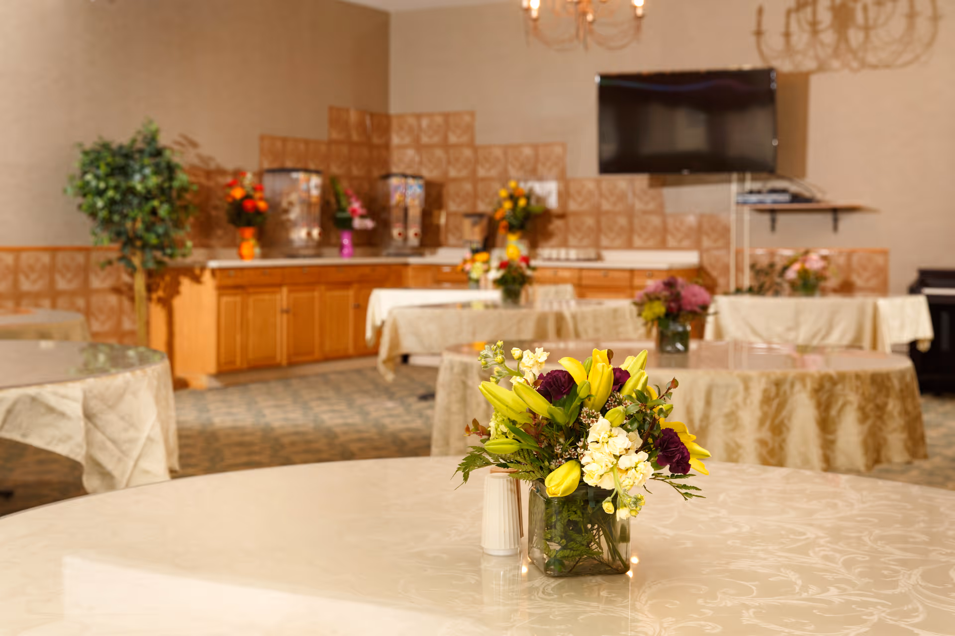 Round tables with tablecloths and floral centerpieces in a banquet-style dining room with a buffet counter and wall-mounted TV.