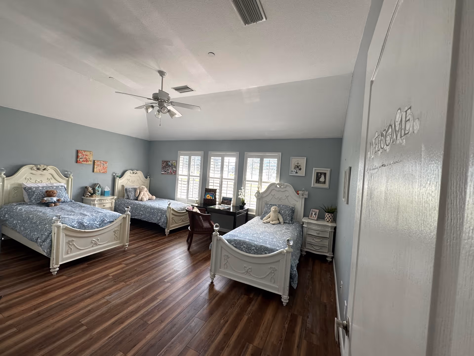 A bedroom with three white ornate twin beds, each with blue patterned bedding and stuffed animals on top. The room has light blue walls, wooden flooring, a ceiling fan with lights, and three windows with white shutters. There are small nightstands beside the beds and framed pictures on the walls.