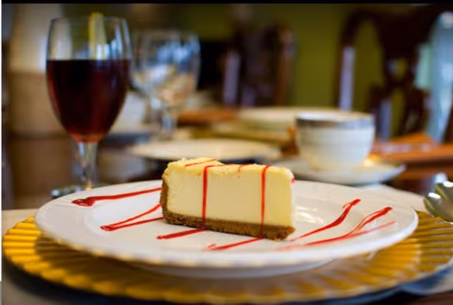 A slice of cheesecake drizzled with red sauce on a white plate, with a glass of red beverage and a cup of coffee in the background on a dining table.
