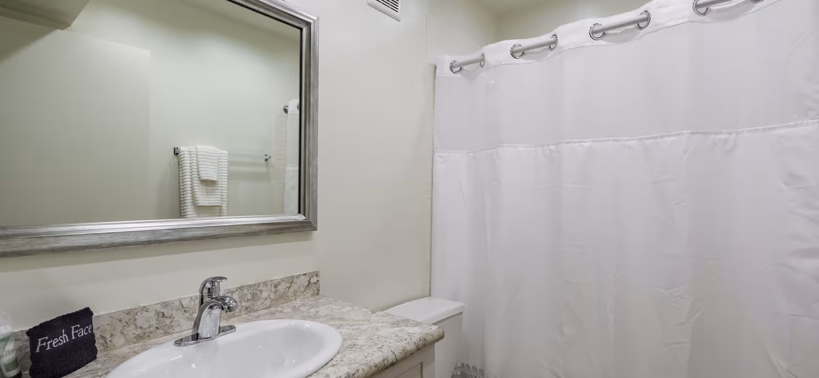 Clean bathroom with a sink on a marble countertop, large framed mirror, towel rack, toilet, and white shower curtain.