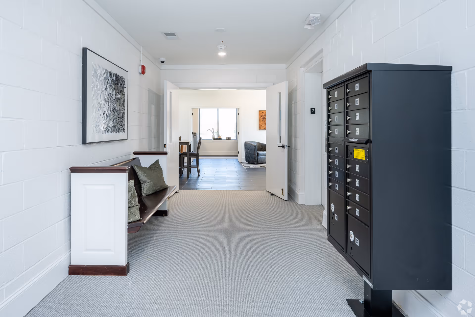 Interior hallway of Serene Haven Apartments with a bench and pillows on the left wall, a set of black mailboxes on the right, and double doors leading to a room with a table, chairs, and a window in the background.