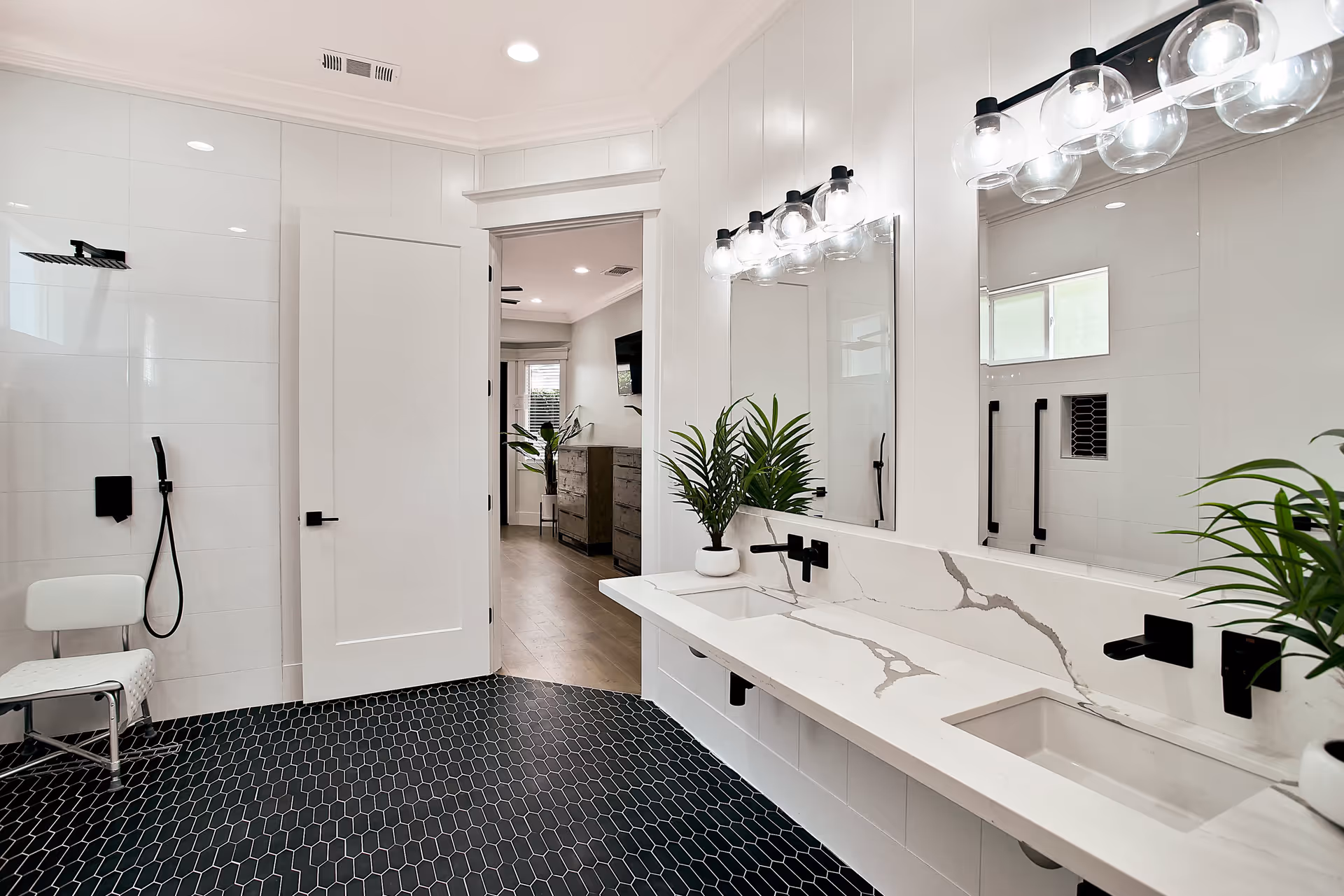 Modern bright bathroom with dual sinks set in a marble countertop, large mirrors, black hexagon tile floor, a walk-in shower and potted plants.