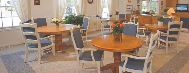 Bright dining room with round wooden tables, cushioned chairs, and floral centerpieces by windows.