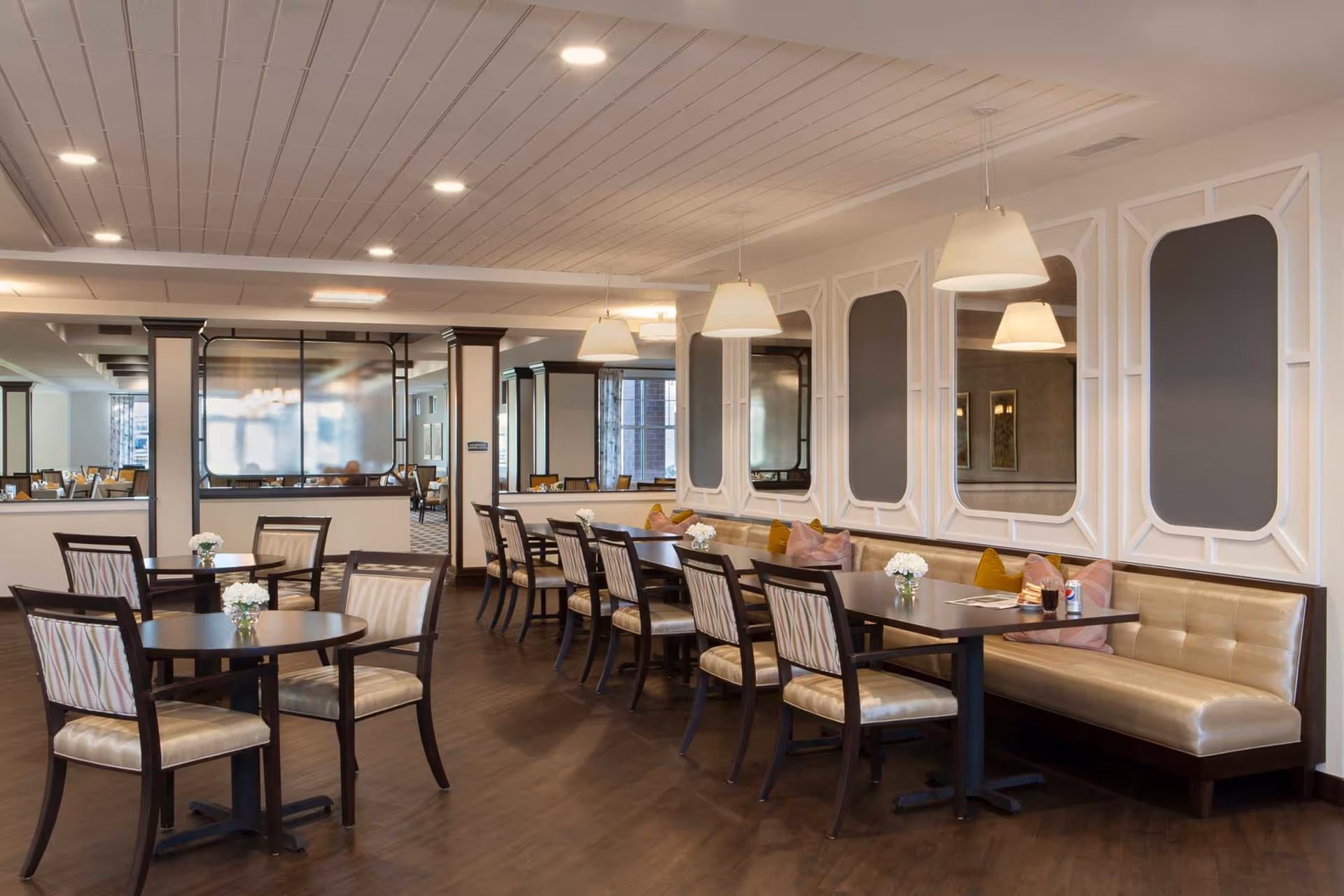 Bright dining room with round and rectangular tables, upholstered chairs and banquette seating beneath pendant lights and decorative wall panels.