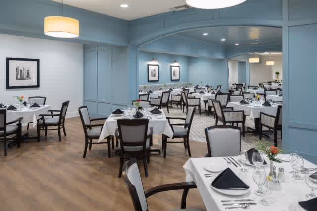 A spacious dining room with multiple tables covered in white tablecloths, each set with black napkins, glassware, and silverware. The room features light blue walls, framed black and white artwork, and modern pendant lighting. The floor is a combination of wood and patterned carpet.