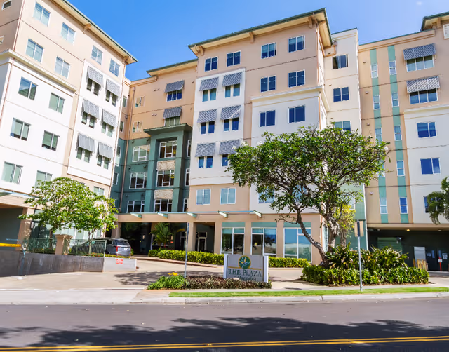 Exterior view of a multi-story senior living facility building with beige and green accents, several windows with awnings, a tree and landscaping in front, and a sign that reads 'The Plaza at Moanalua'.
