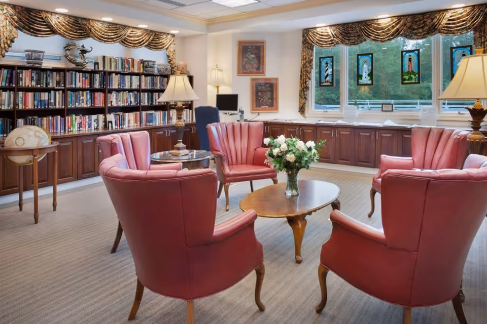 A cozy sitting area in a senior living facility featuring five red leather armchairs arranged around a wooden coffee table with a vase of white flowers. Behind the chairs is a large bookshelf filled with books and decorative items, and a globe on a stand. The room has large windows with patterned valances, stained glass decorations, and two table lamps providing warm lighting.