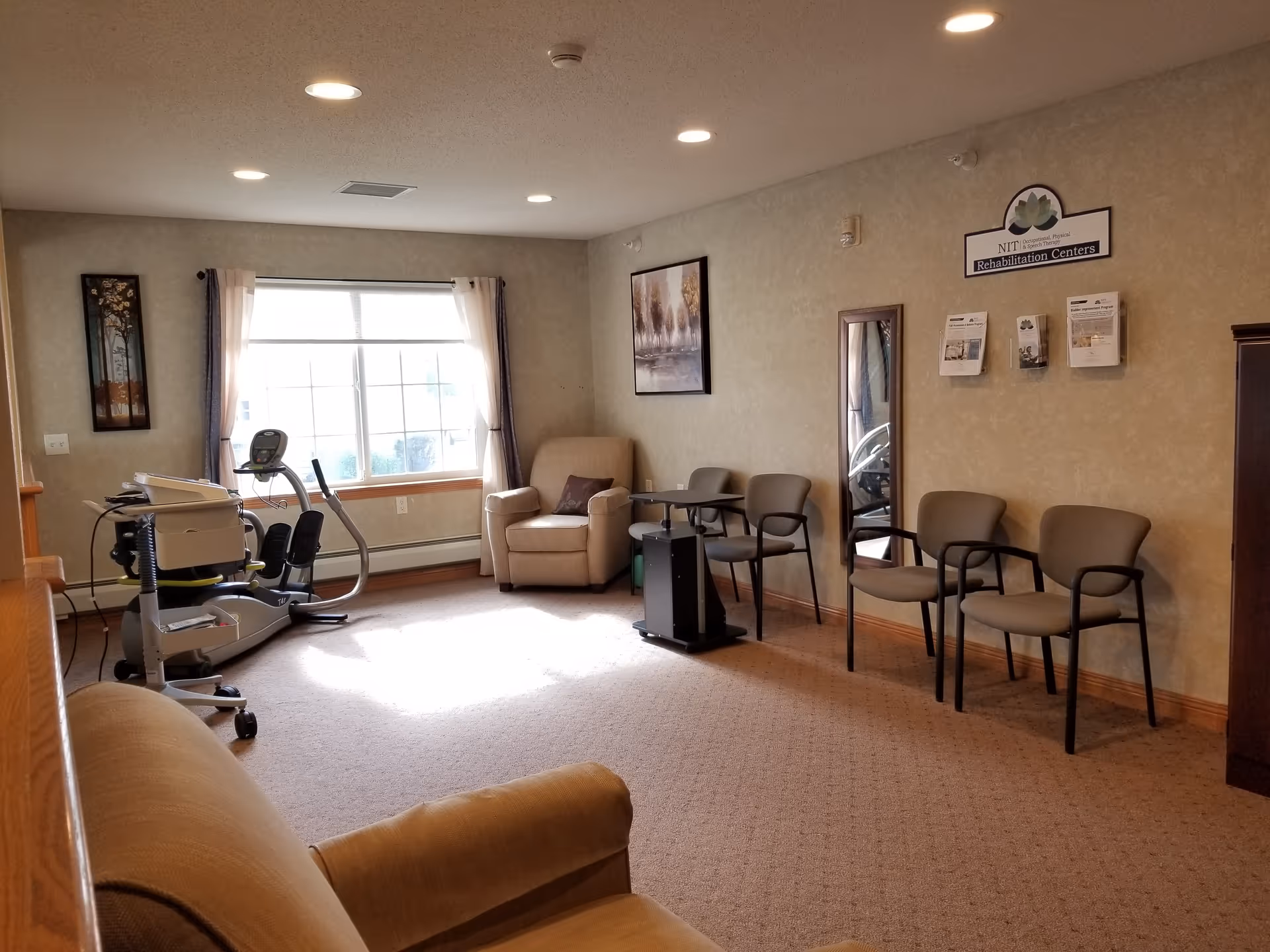 A rehabilitation center room with beige walls and carpet. The room contains a beige armchair, several gray chairs with armrests, a small black table, a mirror on the wall, and exercise equipment near a large window with curtains. There are framed pictures and informational brochures on the walls.