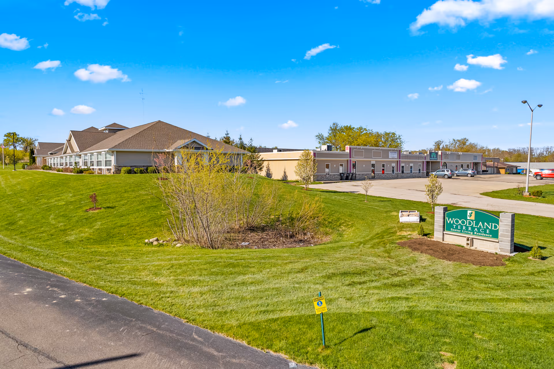 Exterior view of Woodland Terrace of Paw Paw Lake senior living residence on a sunny day with a clear blue sky, green grass, and a parking lot with several cars. The building is a single-story structure with a beige and brown color scheme. A green sign with the facility's name is visible on the lawn near the road.