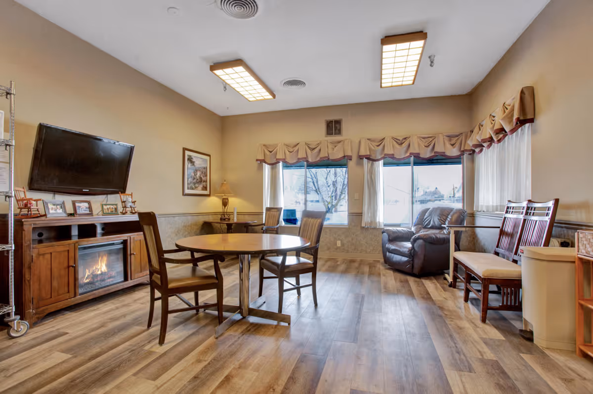 A comfortable common room with a round table and chairs, TV above a fireplace, a recliner and large windows with valances.