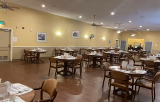 A spacious dining room with multiple round tables set with white tablecloths, napkins, and utensils. Wooden chairs surround each table, and the room has a warm yellow wall color with framed pictures hanging. Ceiling fans and recessed lighting are visible on the ceiling.