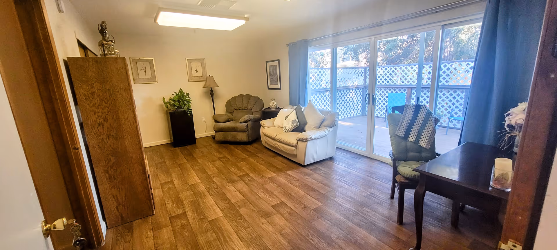 A cozy living room with wooden flooring, a beige loveseat with cushions, a green armchair, a wooden table with a chair draped with a crocheted blanket, a floor lamp, framed artwork on the walls, and a sliding glass door leading to an outdoor patio with lattice fencing.