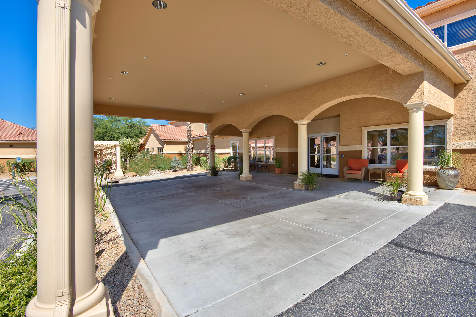 Covered entrance area of Sierra Del Sol Assisted Living with beige columns and arches, outdoor seating with red cushions, potted plants, and a clear blue sky.
