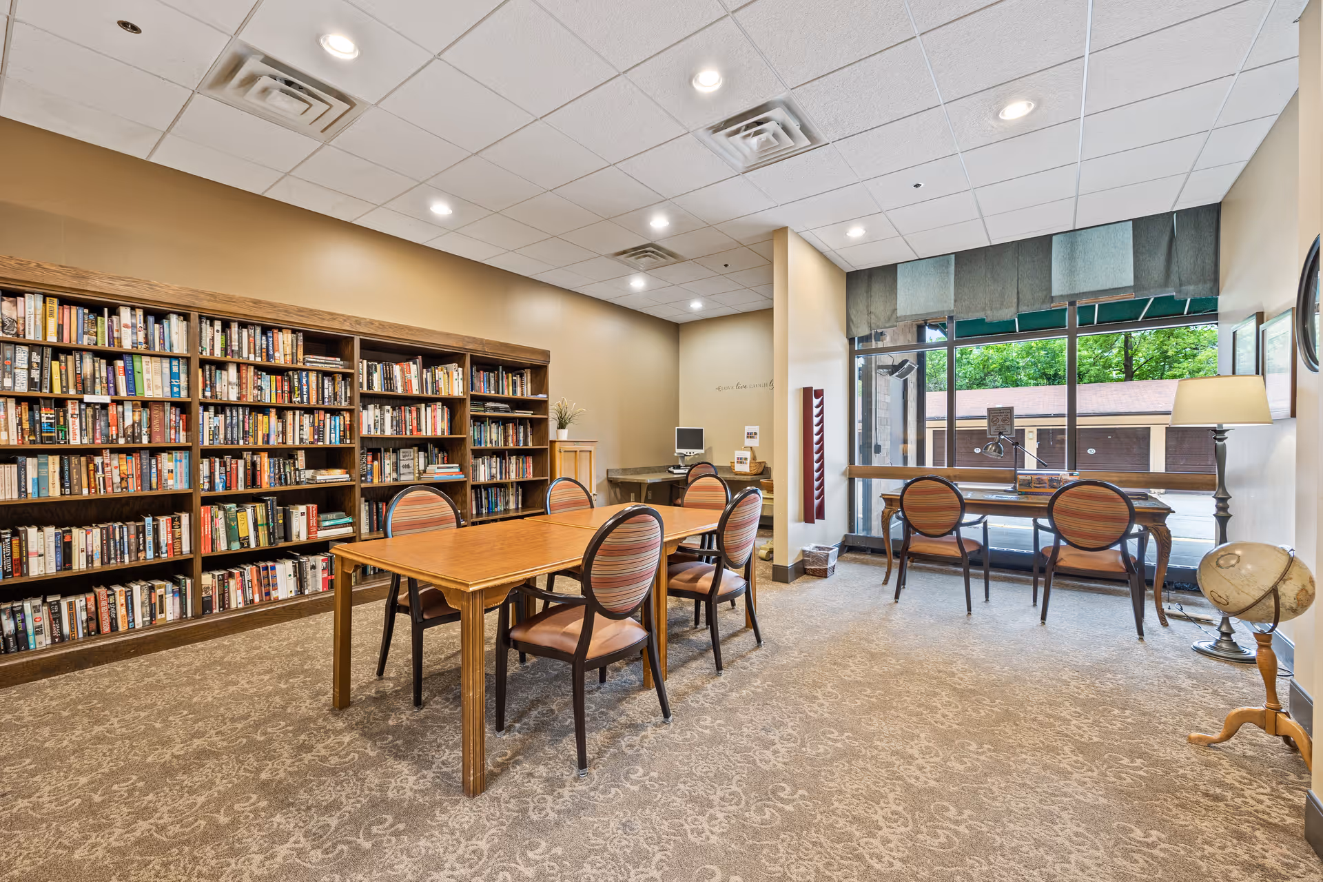 A well-lit senior living facility library room with a large wooden bookshelf filled with books along the left wall. In the center, there is a wooden table surrounded by six chairs with striped upholstery. On the right side, two desks with chairs face a large window with green valances, allowing natural light to fill the room. A floor lamp and a globe on a stand are positioned near the window.