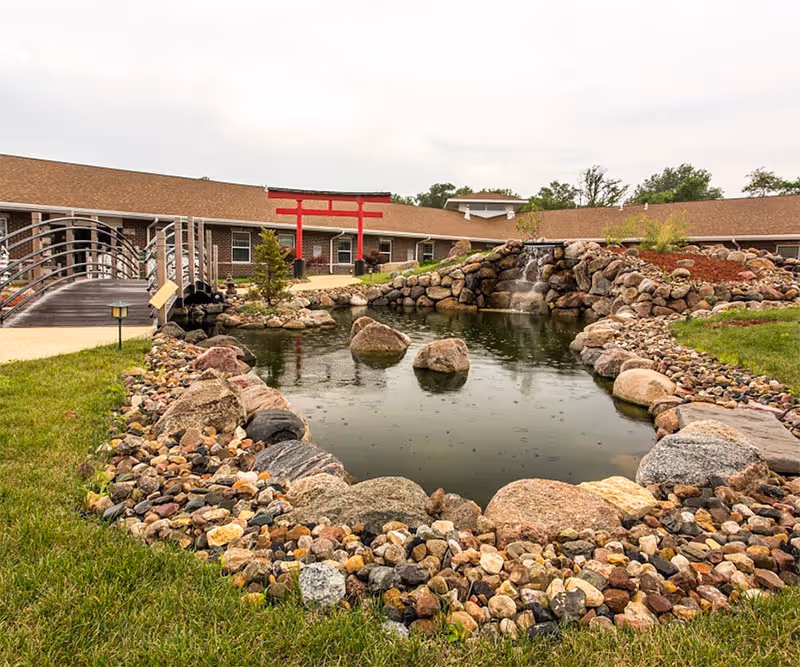 Landscaped courtyard with a rock-bordered pond, small waterfall, footbridge and a red torii-style entrance in front of a single-story building.