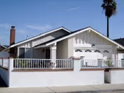 Single-story house with a white exterior, a front porch with a railing, a garage door, a chimney, and a tall palm tree in the background under a clear blue sky.