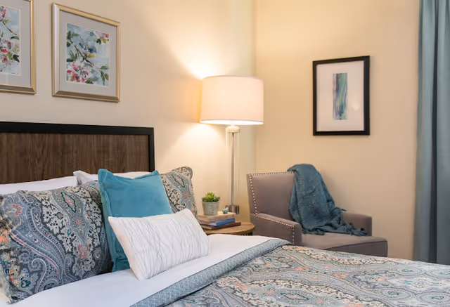 A cozy bedroom with a neatly made bed featuring patterned bedding and multiple pillows. Next to the bed is a wooden nightstand with a small plant, books, and a modern lamp. In the corner, there is a gray armchair draped with a teal throw blanket. The walls are decorated with framed artwork and the room has soft, warm lighting.