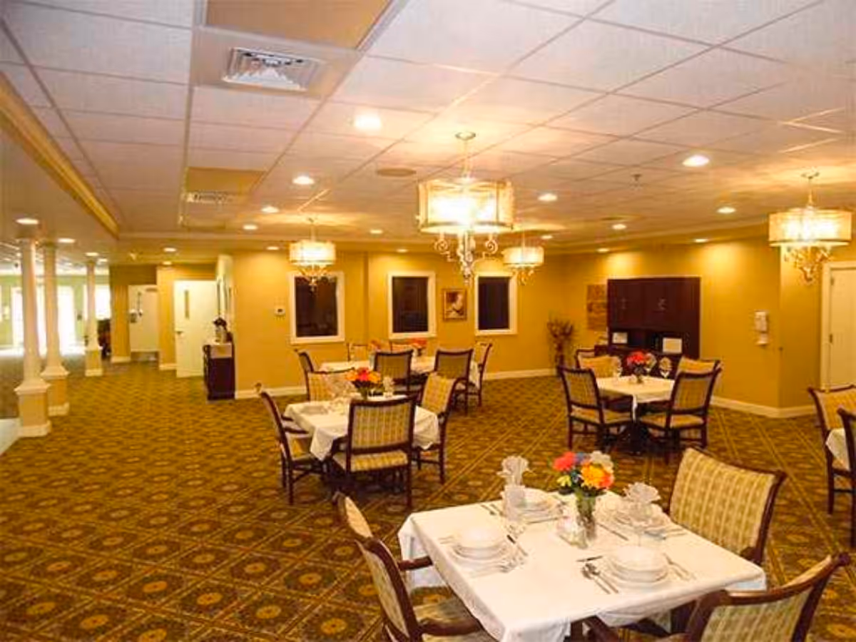 Well-lit dining room with several set tables, upholstered chairs, chandeliers, and patterned carpet.