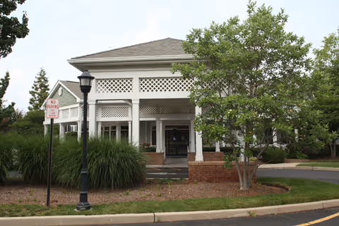 Exterior view of a building with white lattice trim and a covered entrance, surrounded by greenery including trees and bushes, with a streetlamp and a no parking sign in the foreground.