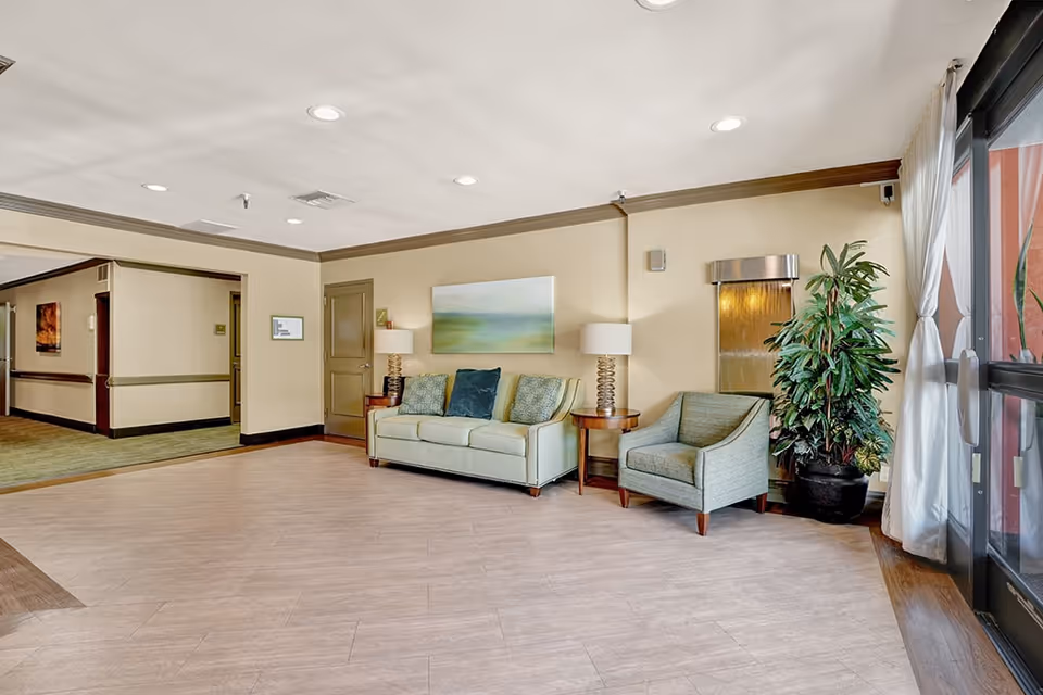 Well-lit seating area in a senior living lobby featuring a sofa, armchair, side tables, artwork, and a potted plant near large windows.