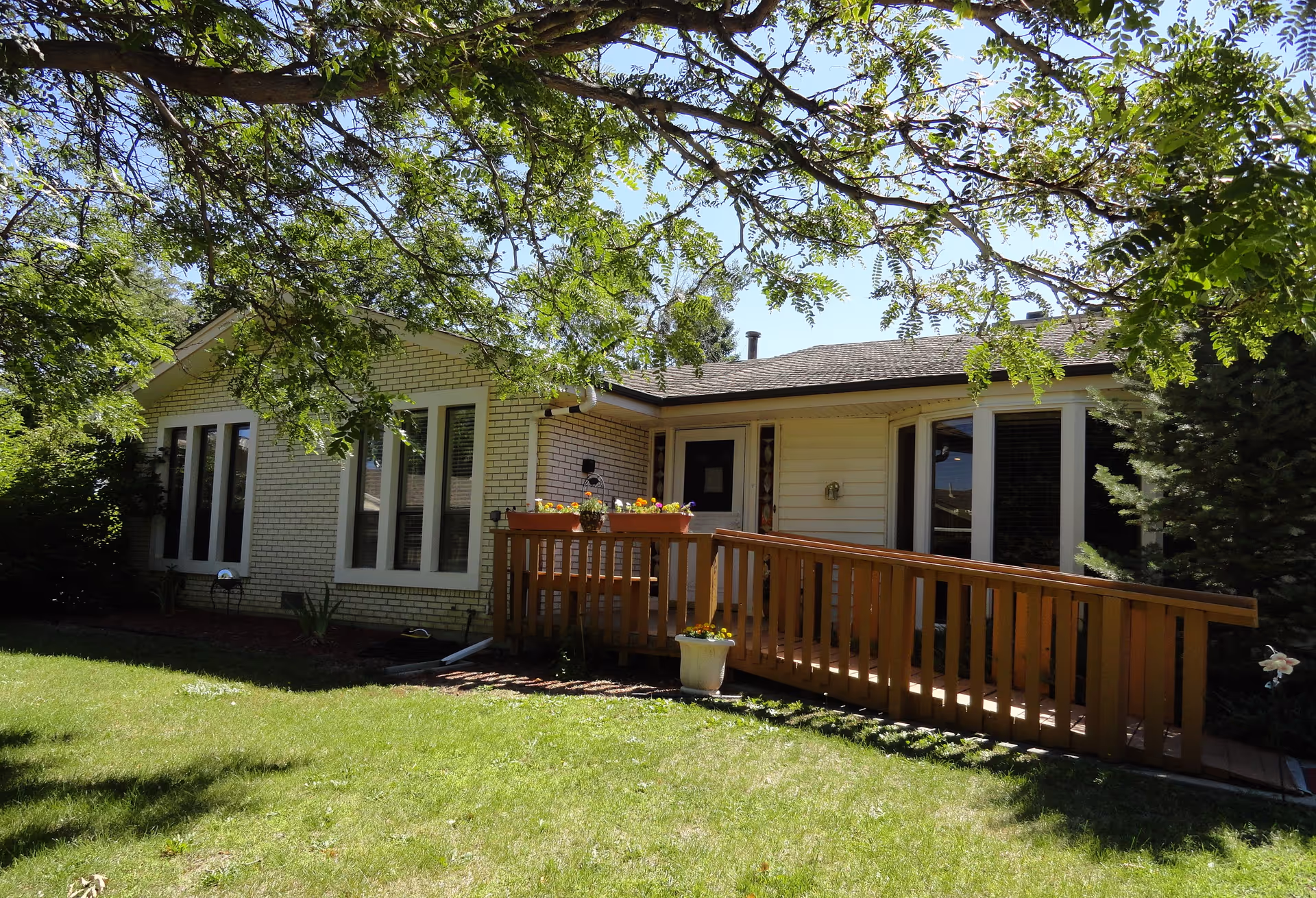 Exterior view of a single-story assisted living facility building with light-colored brick and siding, large windows, a wooden ramp leading to the entrance, and surrounded by green grass and trees.