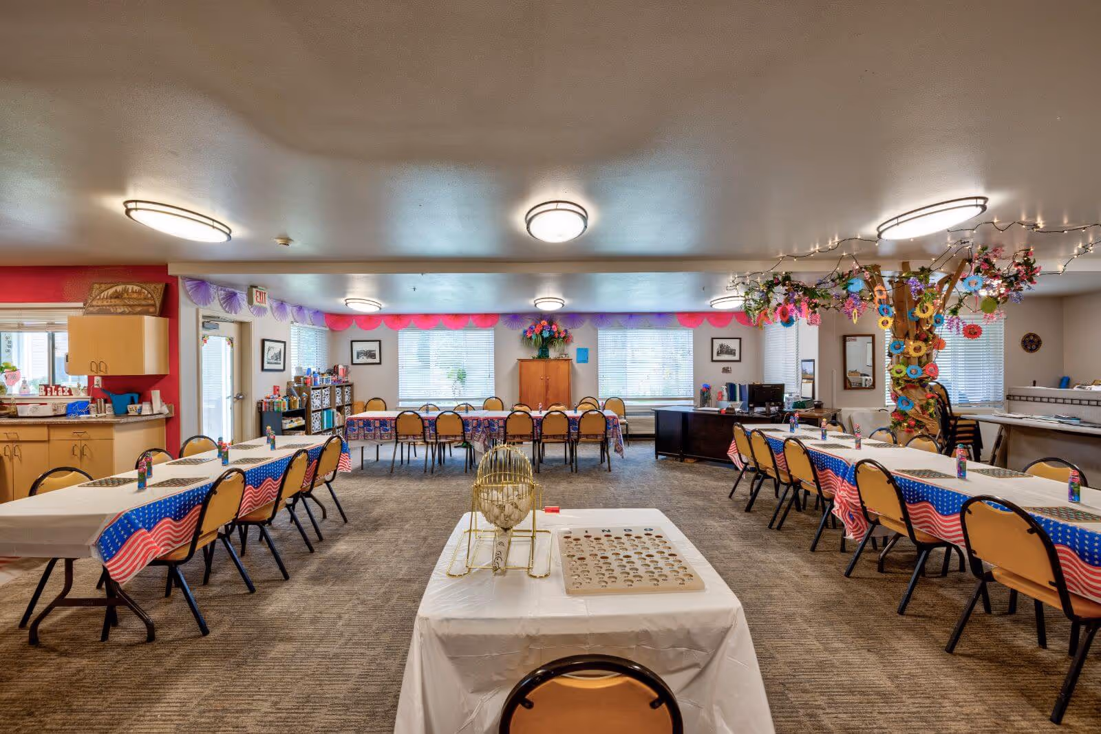 Large decorated communal dining/activity room with rows of tables and chairs, patriotic tablecloths, and a decorated tree-like pillar.
