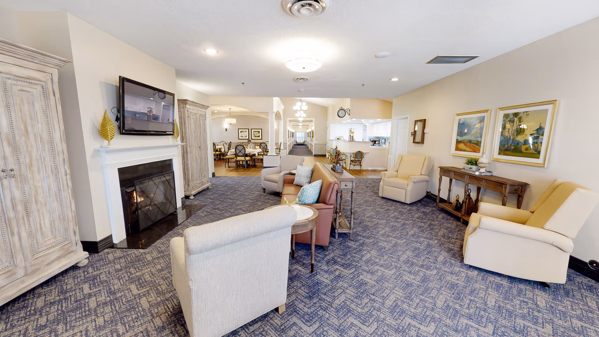 Bright furnished common living area with armchairs, a fireplace and wall-mounted TV, and a dining area visible in the background.