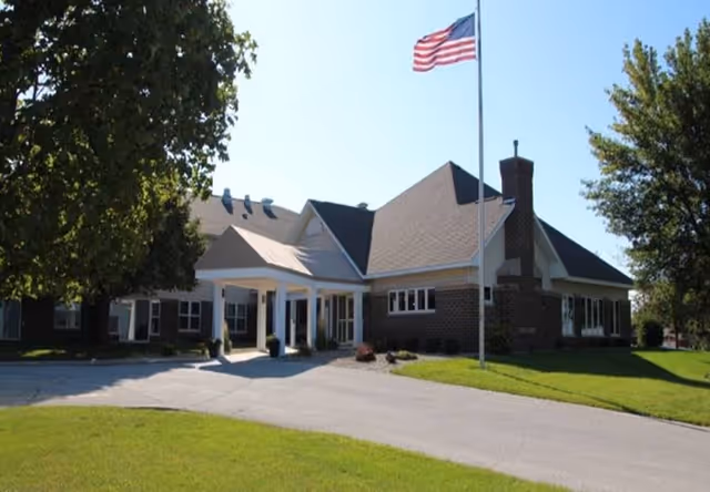 Exterior view of a single-story senior living facility building with a covered entrance, an American flag on a flagpole, and trees on either side under a clear sky.