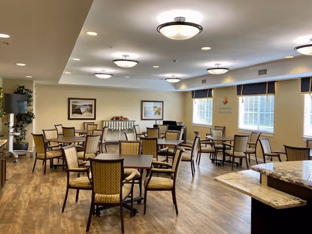 A spacious dining room with multiple square tables and cushioned chairs arranged neatly on a wooden floor. The room has large windows with blinds, framed artwork on the walls, ceiling lights, and a countertop with a granite surface in the foreground.