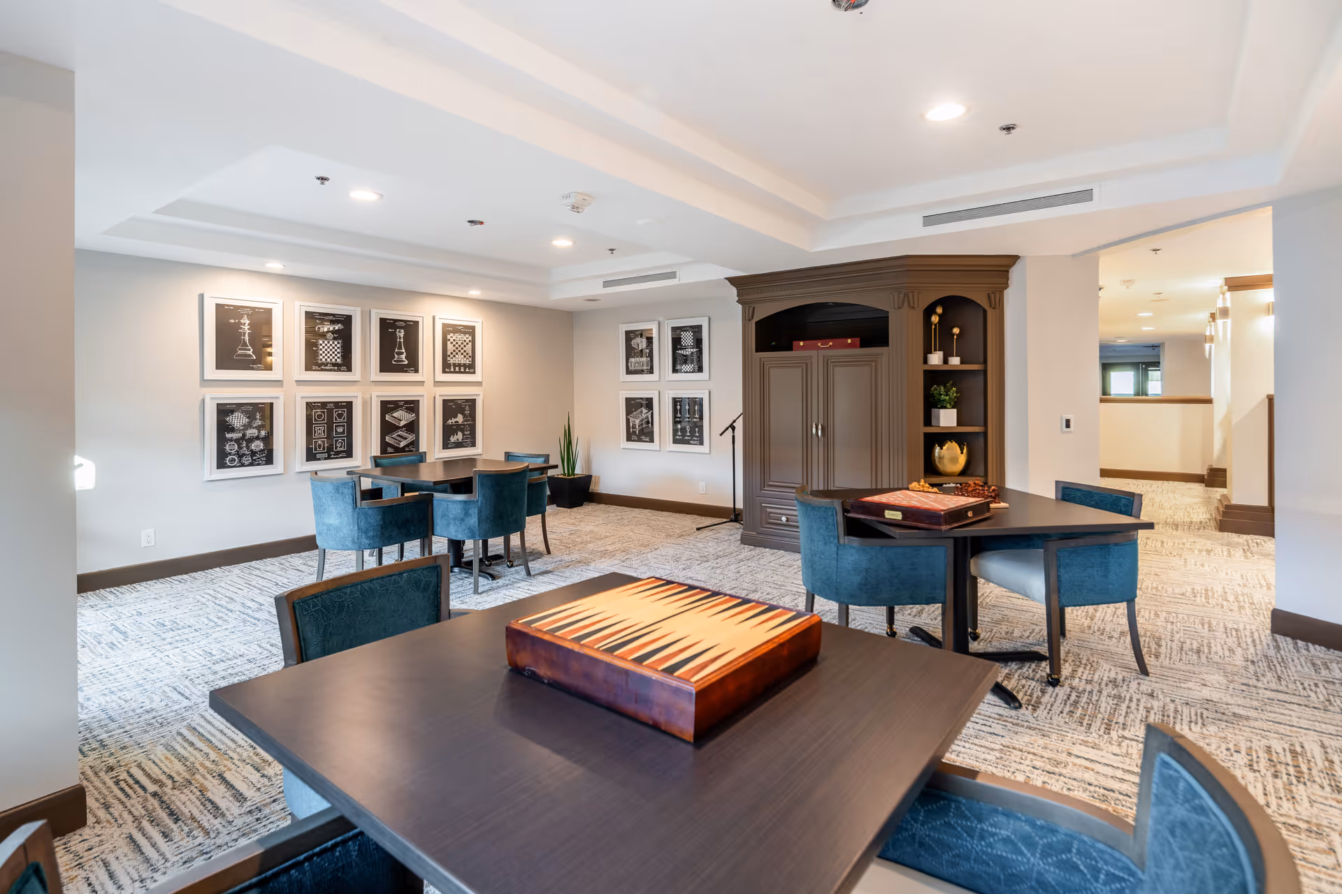 A well-lit common area with several tables and blue upholstered chairs arranged for games. The tables have game boards on them, including a backgammon board in the foreground. The walls feature framed artwork depicting chess and other game-related images. There is a large wooden cabinet with shelves holding decorative items in the background.