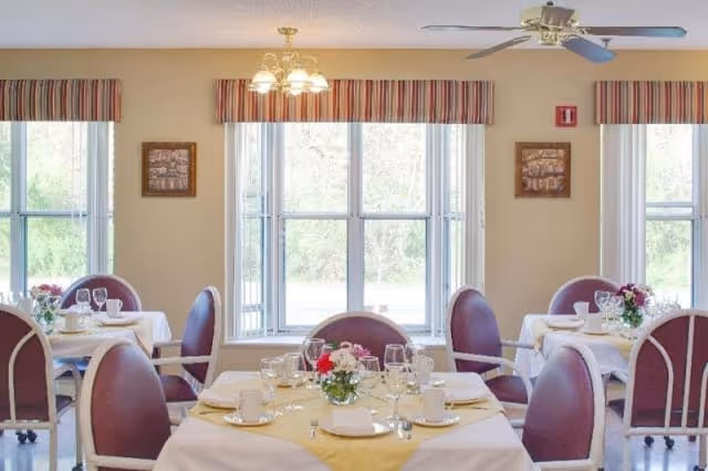 A bright dining room with several round tables covered with white and yellow tablecloths, set with plates, cups, glasses, and floral centerpieces. The room has large windows with striped valances, beige walls, and a ceiling fan with lights.