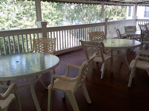 Covered outdoor patio area with multiple round glass-top tables and white plastic chairs arranged around them. The patio has a wooden floor and white railing, with trees and greenery visible in the background.