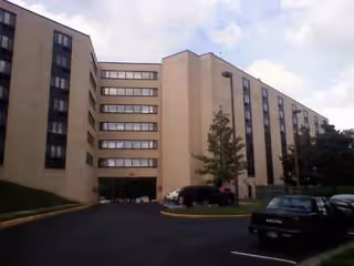 Beige multi-story senior living facility with a parking lot and driveway in view.