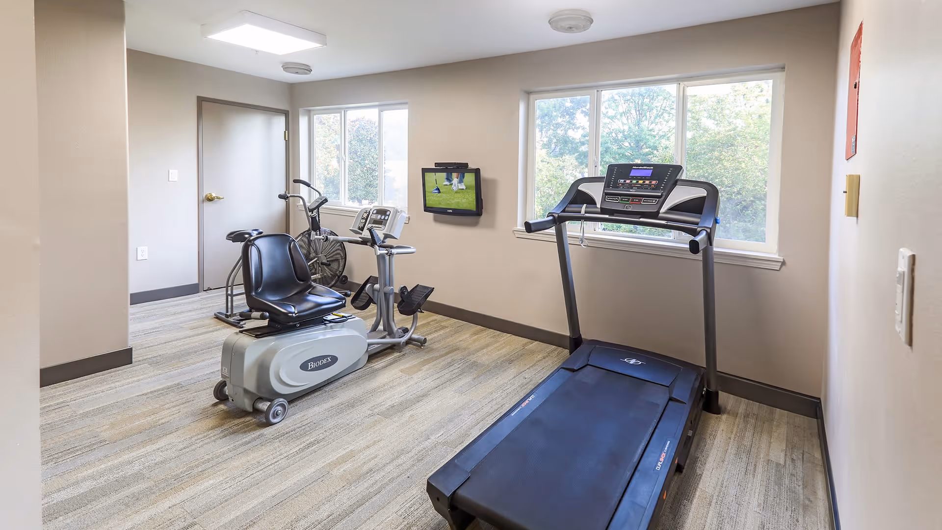 A small exercise room with a treadmill, a recumbent exercise bike, and a stationary bike. The room has two windows letting in natural light, beige walls, and a light-colored carpeted floor. A small TV is mounted on the wall between the windows.