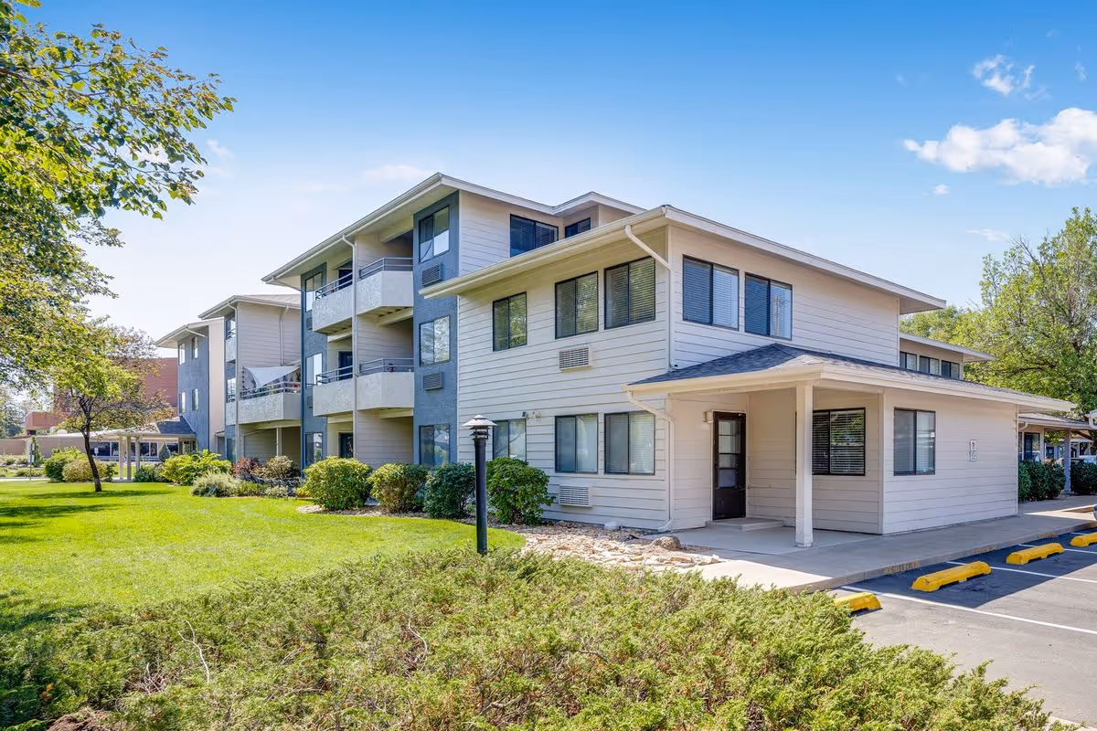 Three-story light-colored senior living building with balconies, landscaped lawn, and parking under a clear blue sky.
