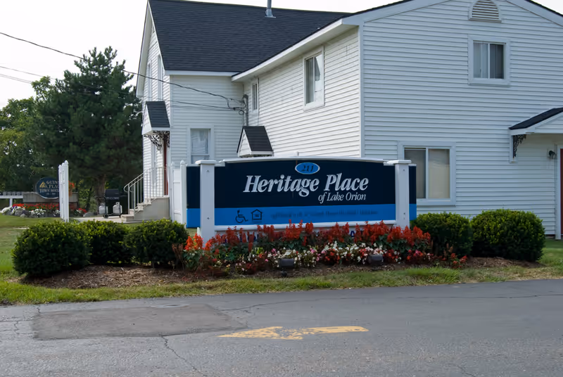 A large sign reading "Heritage Place of Lake Orion" sits in landscaped flower beds in front of a white two-story building.
