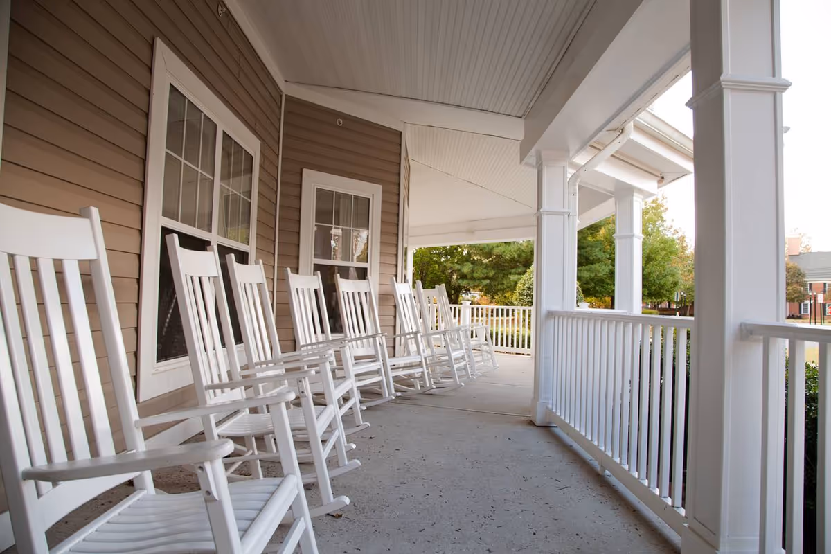 Covered front porch lined with a row of white rocking chairs and a white railing overlooking trees.